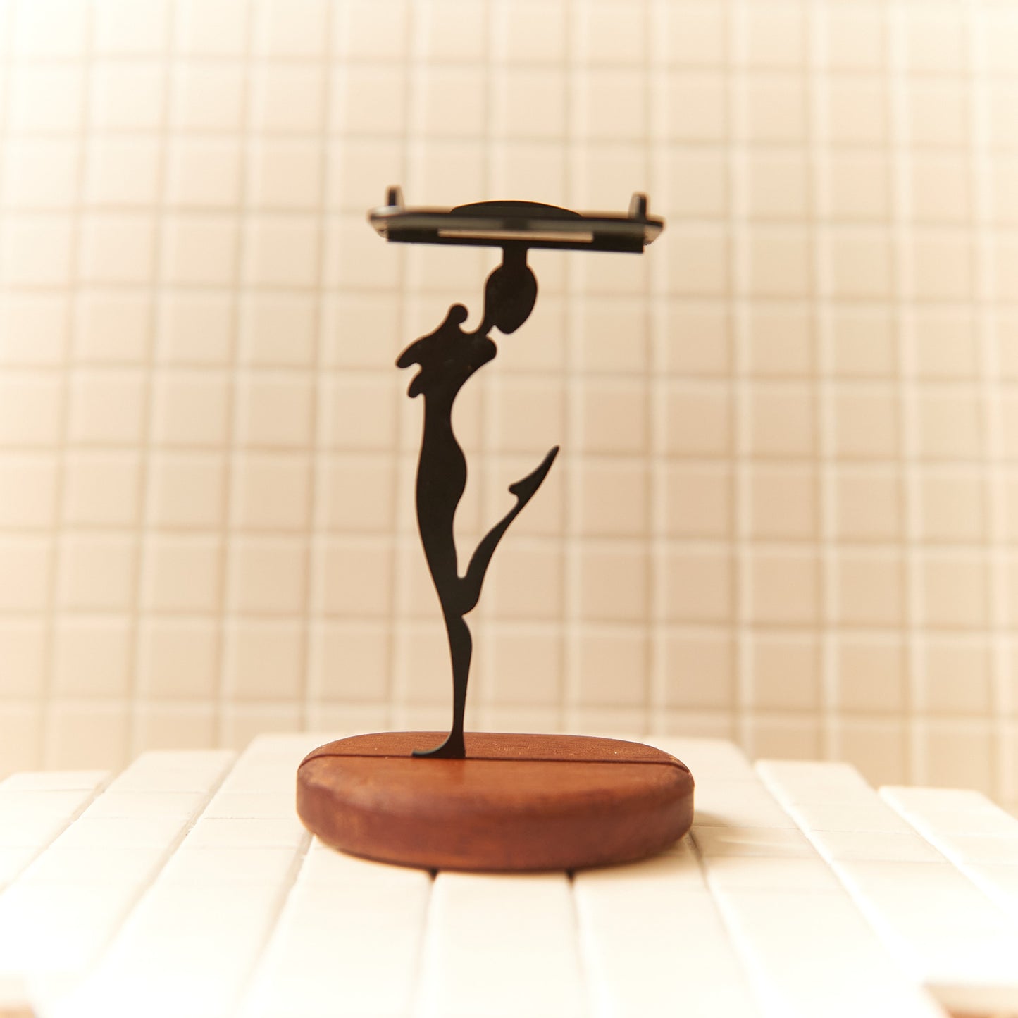 Modern art inspired happy alien sculpture holding a candle plate, designed in black metal with smooth curves and a natural wood base.