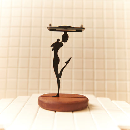 Modern art inspired happy alien sculpture holding a candle plate, designed in black metal with smooth curves and a natural wood base.