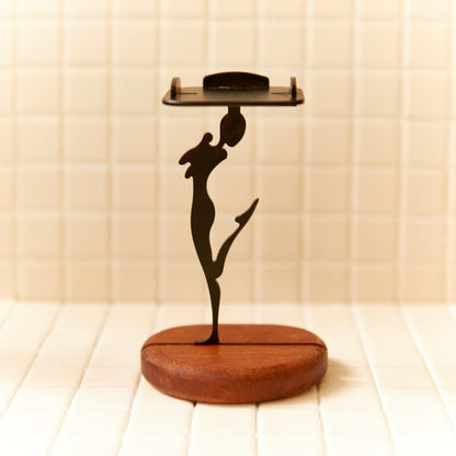 Minimalist black metal alien figurine in a joyful pose, standing on one leg with a raised head, placed on a warm mahogany wooden base — handmade candle holder by Zeugman.